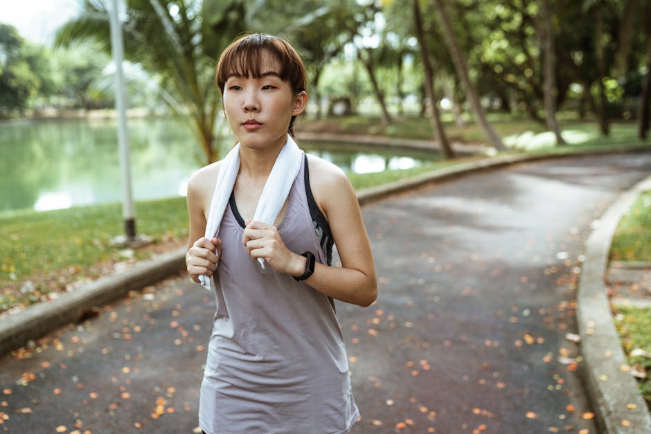 An athletic woman jogging outdoors in a park with a towel around her neck, enhancing wellness